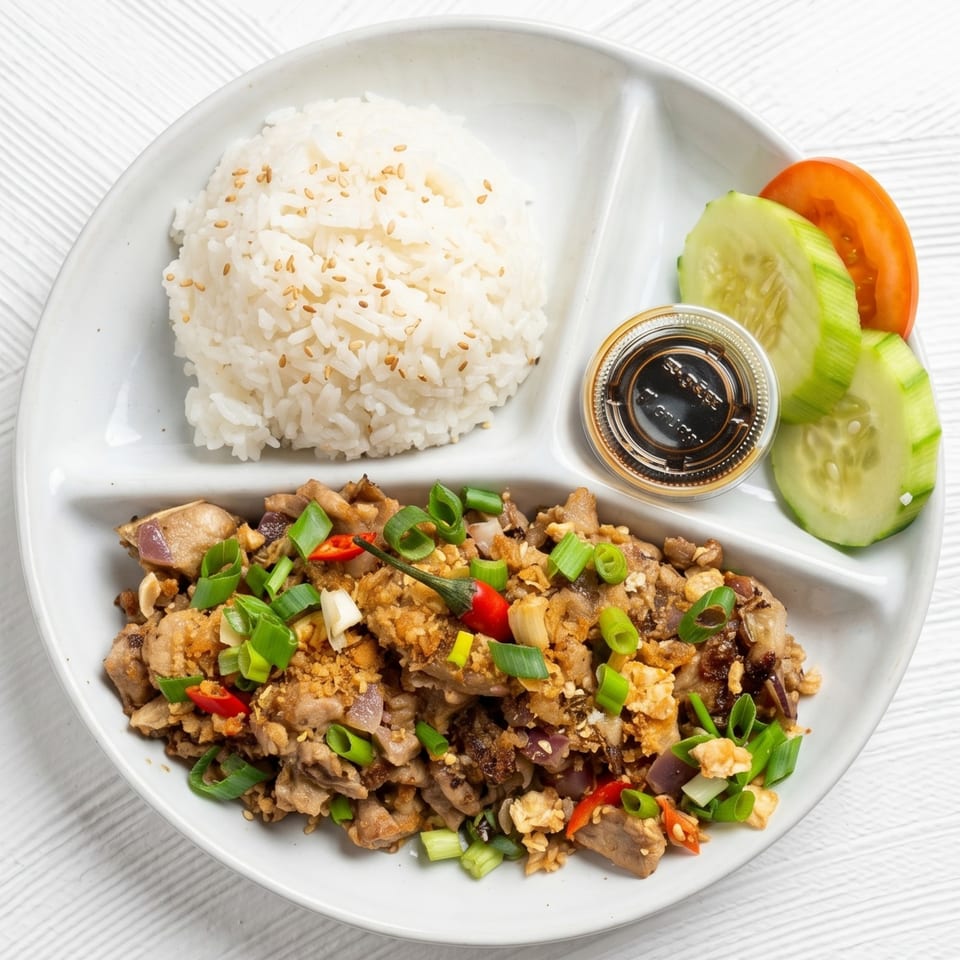 A white divided plate with rice, sisig, cucumber, tomato, and a small cup of sauce.